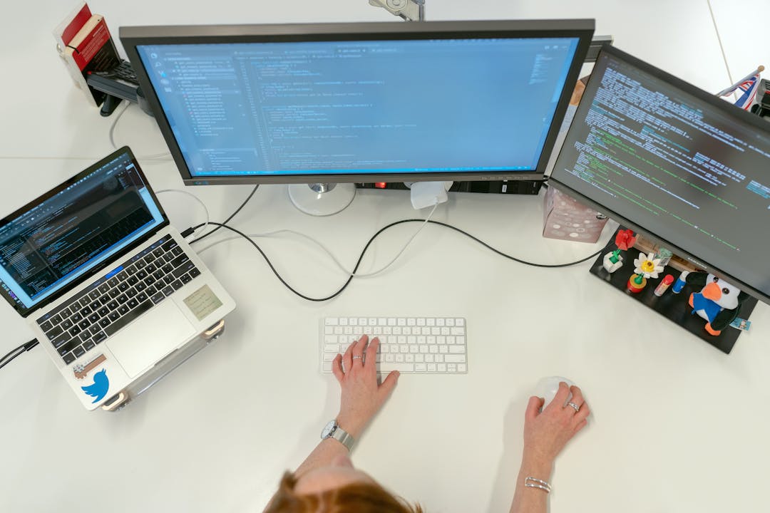 An individual coding at their desk with laptop on the left and two monitors on the right.